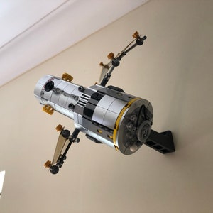 Puede incluir: Un modelo Lego de una estaci&oacute;n espacial de color plateado y negro con detalles dorados, montado en un soporte de pared negro.