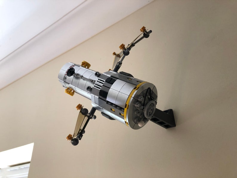 Puede incluir: Un modelo Lego de una estaci&oacute;n espacial de color plateado y negro con detalles dorados, montado en un soporte de pared negro.