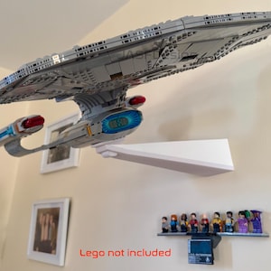 May include: A grey Lego model of a spaceship is mounted on a white shelf. The model has red and blue accents. Below the shelf, a smaller shelf displays Lego figurines. The text "Lego not included" is visible.