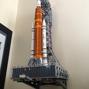May include: A wall mount system for displaying a Lego space shuttle model. The mount is made of black metal and has a gray base. The mount is designed to hold the model securely in place. Lego not included.