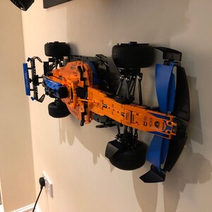 Puede incluir: Un modelo de coche de carreras de F&oacute;rmula 1 Lego negro, naranja y azul montado en una pared. El coche tiene un chasis negro, una carrocer&iacute;a naranja y alas azules.