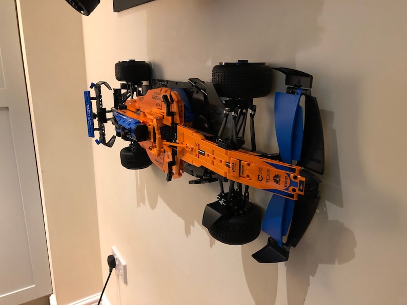 Puede incluir: Un modelo de coche de carreras de F&oacute;rmula 1 Lego negro, naranja y azul montado en una pared. El coche tiene un chasis negro, una carrocer&iacute;a naranja y alas azules.