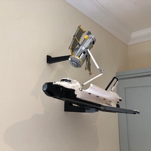 Puede incluir: Un transbordador espacial blanco y negro y una estaci&oacute;n espacial plateada est&aacute;n montados en soportes de pared negros. El transbordador espacial vuela horizontalmente y la estaci&oacute;n espacial est&aacute; inclinada hacia arriba.