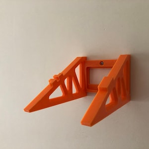 Puede incluir: Un soporte de estante montado en la pared de color naranja. El soporte est&aacute; hecho de pl&aacute;stico y tiene un dise&ntilde;o triangular con una abertura rectangular en el centro. Se fija a una pared blanca con un tornillo.