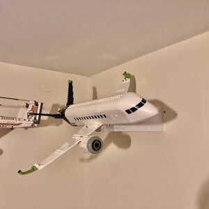 May include: A white airplane model with green accents is mounted on a white wall shelf. The airplane is made of plastic and has a detailed design. The model is displayed in a home setting.