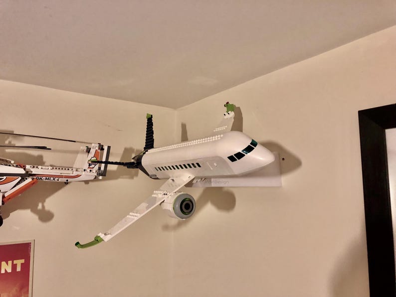 May include: A white airplane model with green accents is mounted on a white wall shelf. The airplane is made of plastic and has a detailed design. The model is displayed in a home setting.