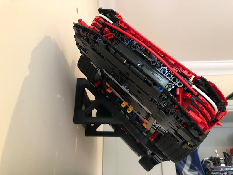 May include: A red and black Lego car model mounted on a black wall mount. The car is upside down and the underside of the car is visible. The car is made of Lego bricks and has a detailed interior.