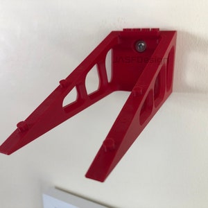 Op de afbeelding: Een rode wandbeugel met een schuin ontwerp. De beugel heeft een reeks uitgesneden vormen en kleine pinnen langs de schuine armen. De tekst "JASFDesign" is zichtbaar op de beugel. Een schroef bevestigt de beugel aan de muur.
