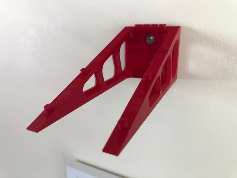 Op de afbeelding: Een rode wandbeugel met een schuin ontwerp. De beugel heeft een reeks uitgesneden vormen en kleine pinnen langs de schuine armen. De tekst "JASFDesign" is zichtbaar op de beugel. Een schroef bevestigt de beugel aan de muur.