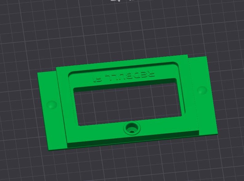 May include: A bright green rectangular plastic component with the text "REDBULL E" embossed on the top. The component has a rectangular cutout in the center and a circular hole at the bottom. Two small circular protrusions are on the sides.