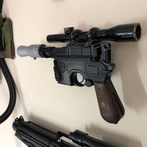 May include: A black and brown prop blaster with a scope, mounted on a wall. The blaster has a silver barrel extension and a brown grip. The scope is black. Other props are visible in the background.