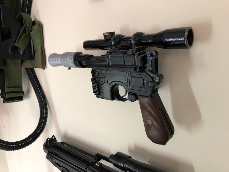May include: A black and brown prop blaster with a scope, mounted on a wall. The blaster has a silver barrel extension and a brown grip. The scope is black. Other props are visible in the background.