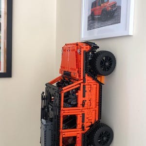 May include: An orange and black Lego model of a Mercedes-Benz G-Wagon, with the vehicle flipped upside down and mounted on a wall. The model is detailed with black wheels and a black interior.