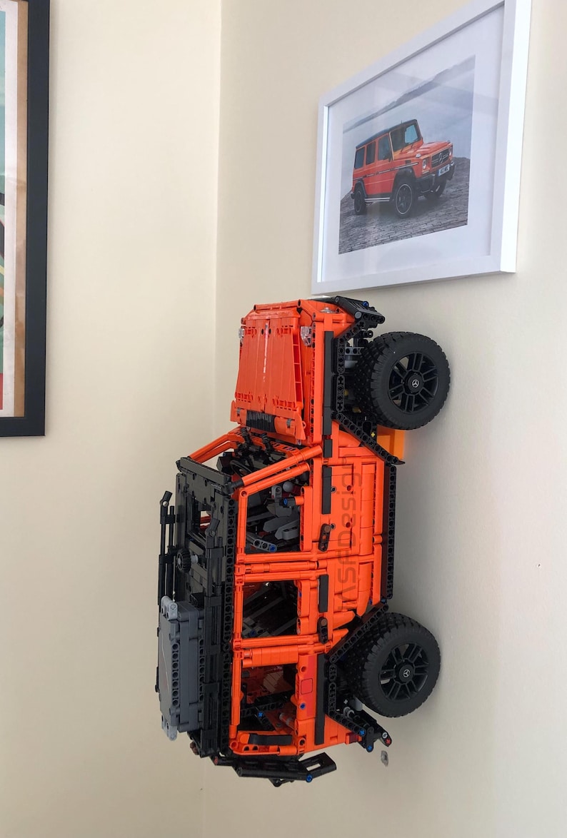 May include: An orange and black Lego model of a Mercedes-Benz G-Wagon, with the vehicle flipped upside down and mounted on a wall. The model is detailed with black wheels and a black interior.