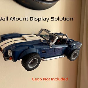 Op de afbeelding: Een blauwe speelgoedauto met witte strepen is aan de muur bevestigd. De auto heeft zwarte banden en wordt weergegeven met de tekst "Wall Mount Display Solution". De tekst "Lego Not Included" is ook zichtbaar.