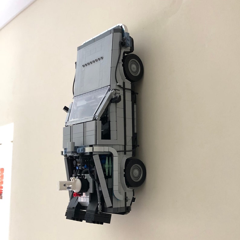 May include: A gray and white Lego model of the DeLorean time machine from the Back to the Future movie. The model is mounted on a wall and is facing forward. The car has a detailed interior and exterior, including the iconic flux capacitor.