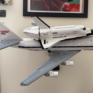Op de afbeelding: Een wit LEGO-model van een NASA-spaceshuttle bovenop een Boeing 747-vliegtuig. Het model heeft het NASA-logo en de tekst "United States". Het vliegtuig heeft de tekst "JASEDesign" op de zijkant.