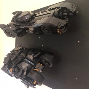 May include: Two black Lego Batmobiles, one with a sleek, futuristic design and the other with a more rugged, armored look, are displayed on a black background.