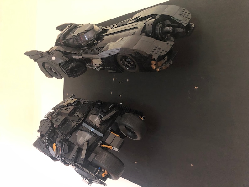 May include: Two black Lego Batmobiles, one with a sleek, futuristic design and the other with a more rugged, armored look, are displayed on a black background.