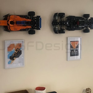 May include: Two Lego Formula 1 race cars are mounted on a wall, one orange and blue and the other black.  Two framed posters of Formula 1 cars are also on the wall.