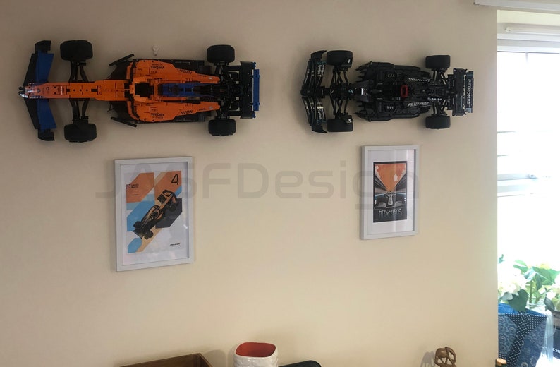 May include: Two Lego Formula 1 race cars are mounted on a wall, one orange and blue and the other black.  Two framed posters of Formula 1 cars are also on the wall.