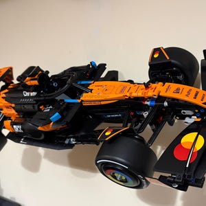 May include: A black and orange Lego McLaren Formula 1 race car model. The model features black tires, blue accents, and various sponsor logos. The car is mounted on a wall, showcasing its aerodynamic design and intricate details.