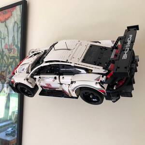 May include: A white Lego Porsche 911 RSR race car with red and black accents. The car is displayed on a wall mount with the rear spoiler raised. The car has the word "Porsche" printed on the spoiler.
