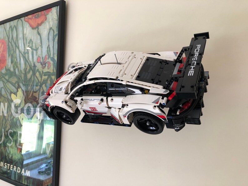 May include: A white Lego Porsche 911 RSR race car with red and black accents. The car is displayed on a wall mount with the rear spoiler raised. The car has the word "Porsche" printed on the spoiler.