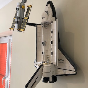 May include: A white and black NASA Discovery space shuttle model with open cargo bay doors, connected to a satellite model with yellow solar panels. The models are mounted on a beige wall.