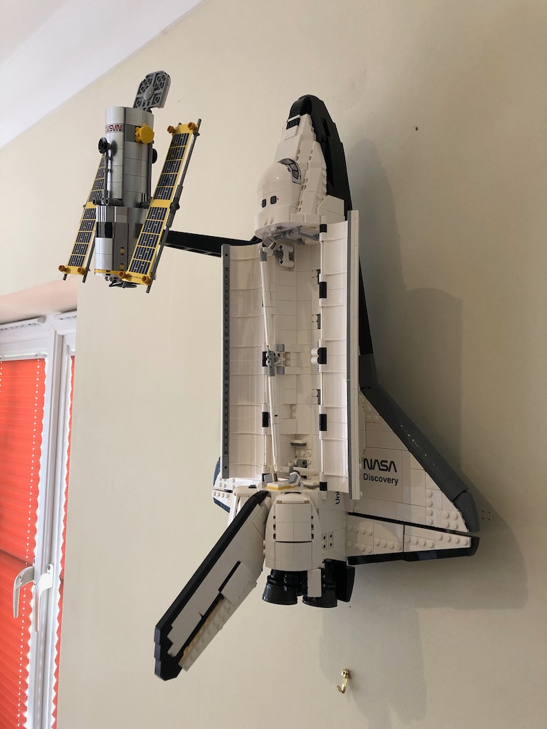 May include: A white and black NASA Discovery space shuttle model with open cargo bay doors, connected to a satellite model with yellow solar panels. The models are mounted on a beige wall.