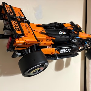 May include: An orange and black race car model with the text "ACCELERA" and "OKX" printed on the body. The model has large black tires and is mounted on a wall. The model also has the text "Chrome" and "Dell" printed on the body.