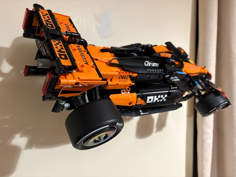 May include: An orange and black race car model with the text "ACCELERA" and "OKX" printed on the body. The model has large black tires and is mounted on a wall. The model also has the text "Chrome" and "Dell" printed on the body.