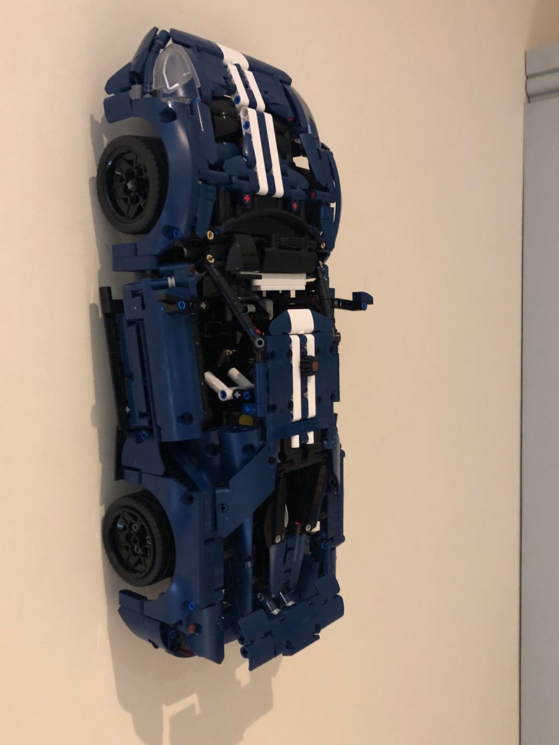 May include: A blue Lego model of a Ford GT race car with white racing stripes. The car is displayed on a wall with the wheels facing out.