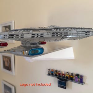 Puede incluir: Un modelo gris de una nave espacial, posiblemente la Enterprise de Star Trek, est&aacute; montado en una estanter&iacute;a blanca. El modelo tiene detalles intrincados y se exhibe contra una pared neutra. Debajo hay una peque&ntilde;a colecci&oacute;n de figuritas.