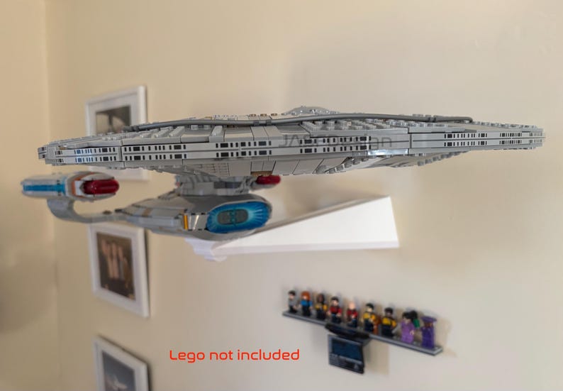Puede incluir: Un modelo gris de una nave espacial, posiblemente la Enterprise de Star Trek, est&aacute; montado en una estanter&iacute;a blanca. El modelo tiene detalles intrincados y se exhibe contra una pared neutra. Debajo hay una peque&ntilde;a colecci&oacute;n de figuritas.