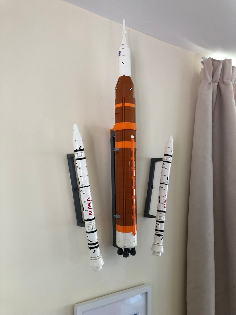 May include: Three white and orange Lego rockets mounted on a wall with the word "NASA" printed on the side of each rocket.