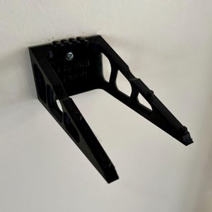 May include: A black, angled wall mount, attached to a white wall. The mount has a rectangular base with a series of holes and a screw. The angled arms extend outward, with a textured surface on the ends. The text "BRYAN 2004" is visible.