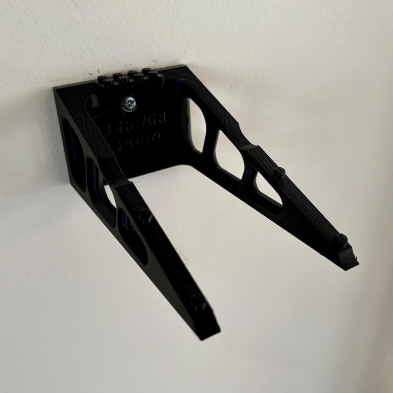May include: A black, angled wall mount, attached to a white wall. The mount has a rectangular base with a series of holes and a screw. The angled arms extend outward, with a textured surface on the ends. The text "BRYAN 2004" is visible.