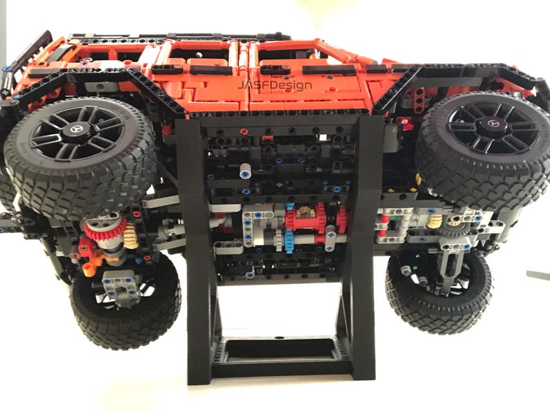 May include: A black and orange Lego model of a Mercedes-Benz G-Wagon on a black display stand. The model is upside down, showing the underside of the vehicle.