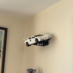 May include: A white model car, possibly a Lamborghini Countach, is mounted on a black wall shelf. The car is facing the right side of the image and is angled slightly upwards. The shelf is attached to the wall with screws.