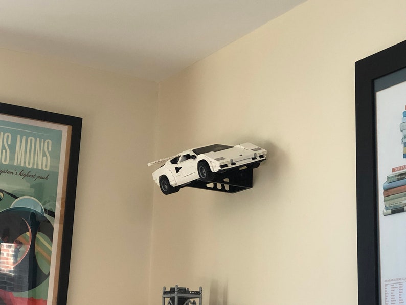 May include: A white model car, possibly a Lamborghini Countach, is mounted on a black wall shelf. The car is facing the right side of the image and is angled slightly upwards. The shelf is attached to the wall with screws.