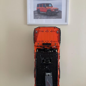 May include: A black and orange Lego model of a Mercedes-Benz G-Wagon, with a picture of the real car in a white frame above it.