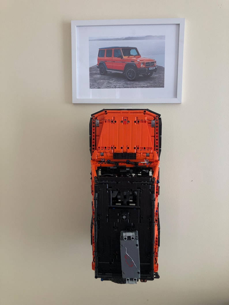 May include: A black and orange Lego model of a Mercedes-Benz G-Wagon, with a picture of the real car in a white frame above it.