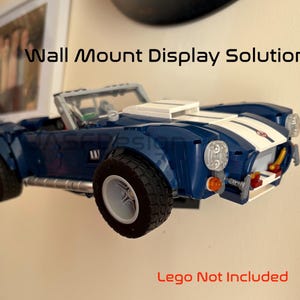 Op de afbeelding: Een blauwe en witte speelgoedauto aan de muur gemonteerd. De auto heeft zwarte banden en een witte streep in het midden. Tekst op de afbeelding luidt "Wall Mount Display Solution" en "Lego Not Included".