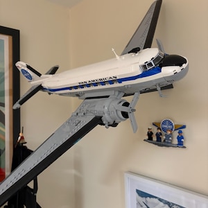 May include: A white and blue Pan American airplane model with gray wings and propellers. The model is mounted on a wall, with the text "PAN AMERICAN" visible on the side. A small Pan Am logo and figurines are also on the wall.
