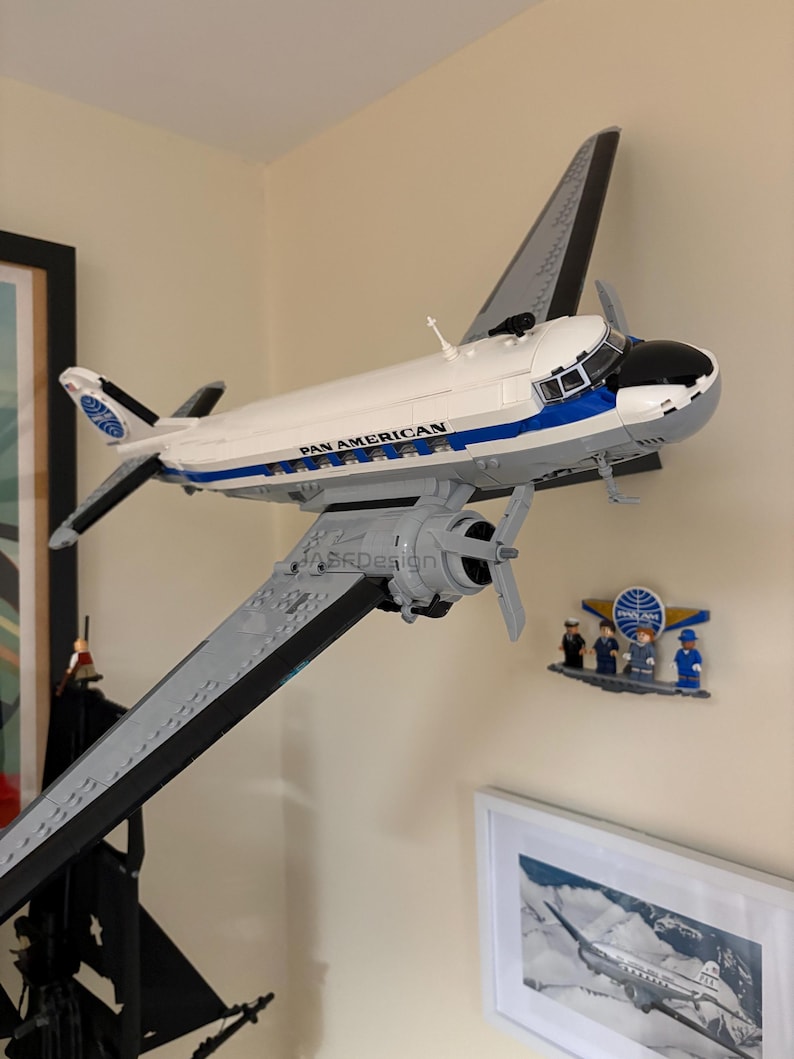 May include: A white and blue Pan American airplane model with gray wings and propellers. The model is mounted on a wall, with the text "PAN AMERICAN" visible on the side. A small Pan Am logo and figurines are also on the wall.