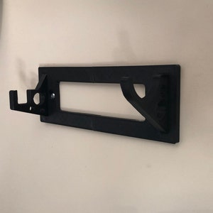 May include: Black plastic wall mount with two hooks and a rectangular cutout. The mount is designed to hold a tool or other object.