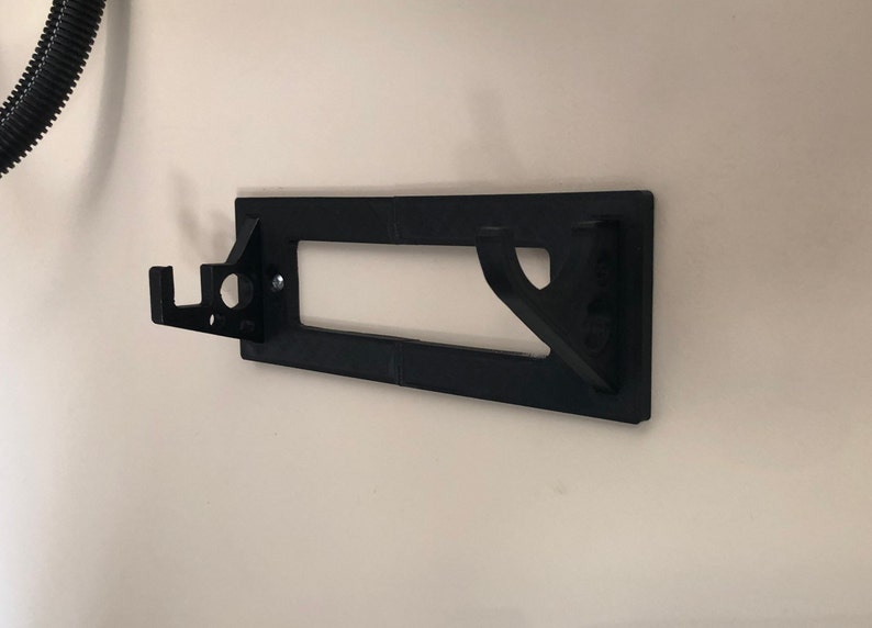 May include: Black plastic wall mount with two hooks and a rectangular cutout. The mount is designed to hold a tool or other object.