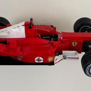 May include: A red and white Lego Ferrari Formula 1 race car model. The car features black tires with white lettering, and sponsor logos including Shell, Bridgestone, and Vodafone. The car has a prominent rear wing.
