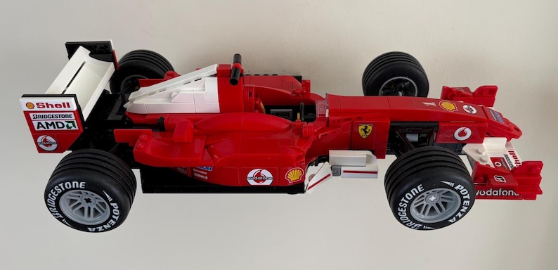 May include: A red and white Lego Ferrari Formula 1 race car model. The car features black tires with white lettering, and sponsor logos including Shell, Bridgestone, and Vodafone. The car has a prominent rear wing.